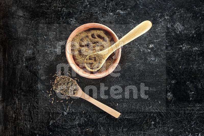 A wooden bowl and 2 wooden spoons full of cumin powder and cumin seeds on a textured black flooring