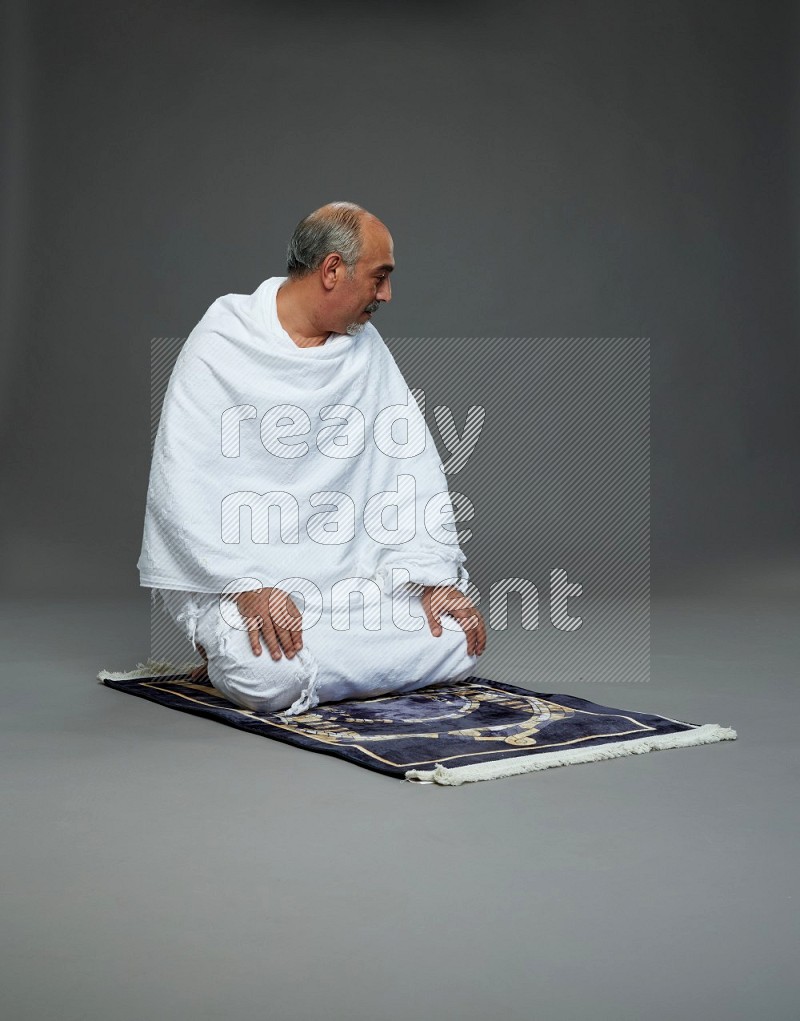 A man wearing Ehram sitting on prayer mat on gray background