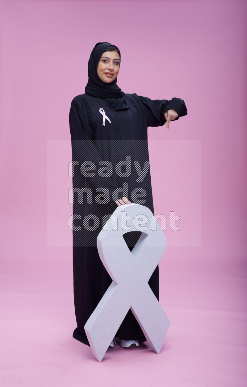 Saudi woman wearing pink ribbon on abaya standing holding awareness ribbon on plain pink background