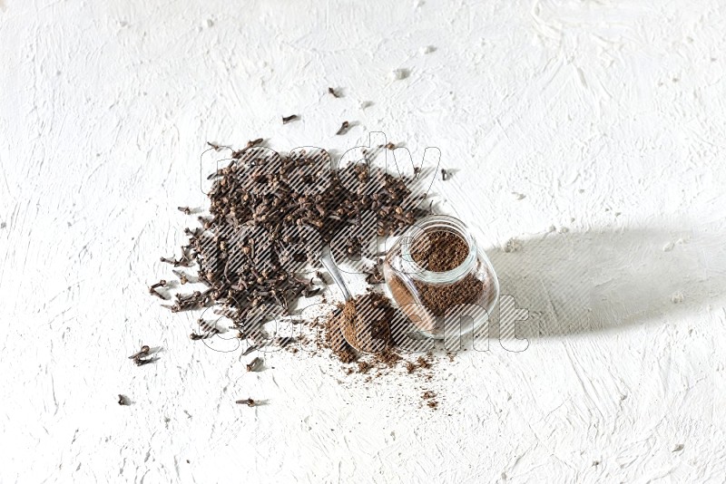 A glass spice jar and a metal spoon full of cloves powder and cloves spread on textured white flooring