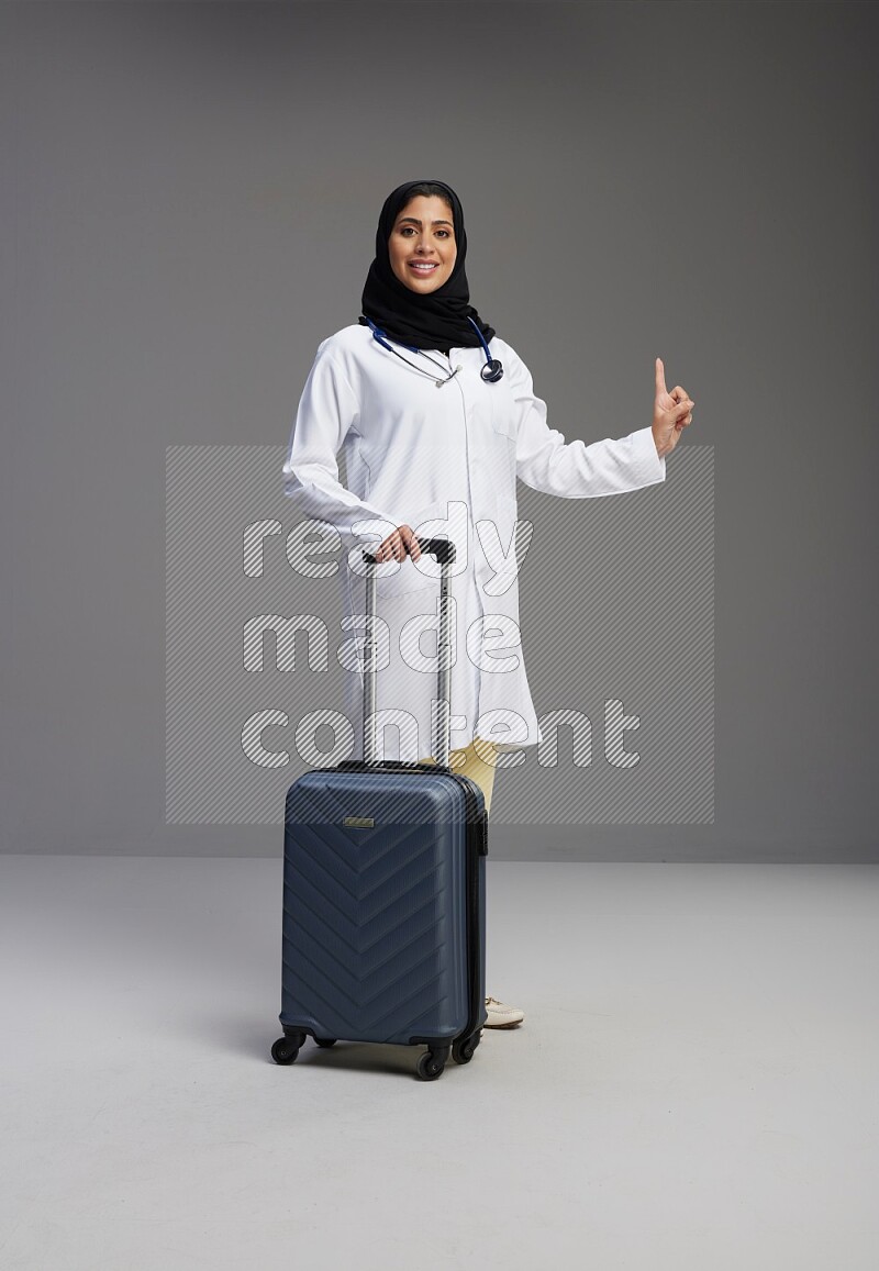 Saudi woman wearing lab coat with stethoscope standing holding Travel bag on Gray background