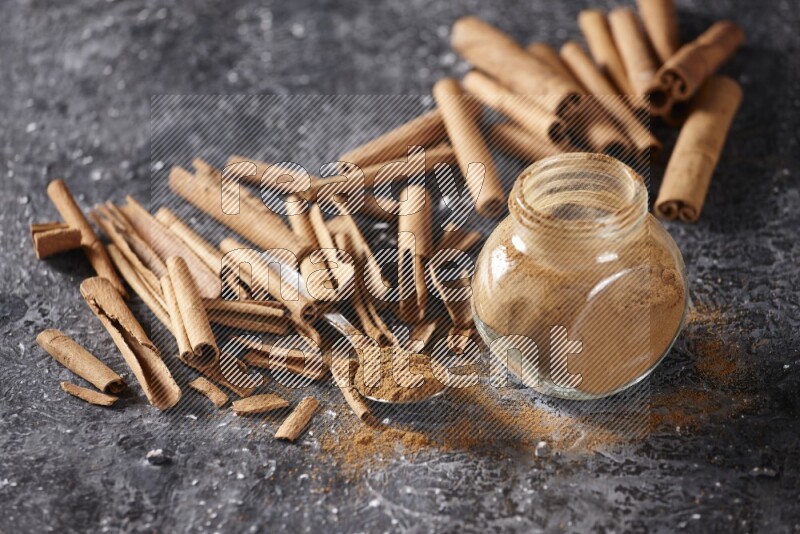 Herbal glass jar and a metal spoon full of cinnamon powder surrounded by cinnamon sticks on textured black background