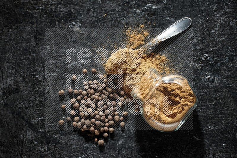 A flipped glass spice jar and metal spoon full of allspice powder and powder spilled out of it with whole balls on a textured black flooring