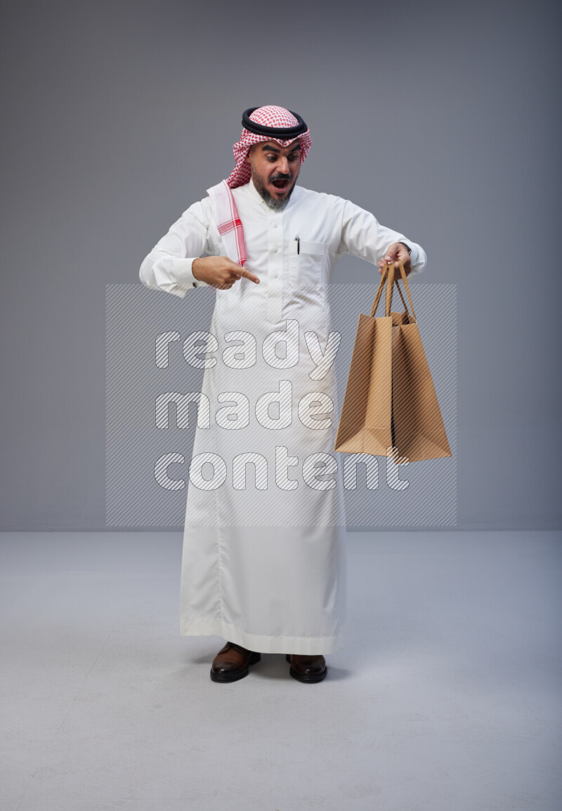 Saudi man Wearing Thob and red Shomag standing holding shopping bag on Gray background