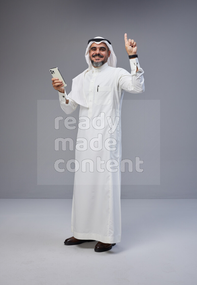 Saudi man Wearing Thob and white Shomag standing texting on phone on Gray background