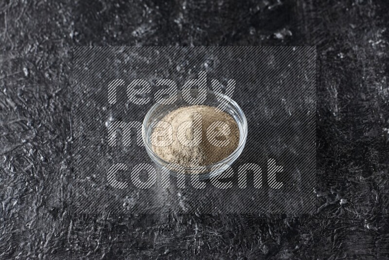A glass bowl full of white pepper powder on textured black flooring