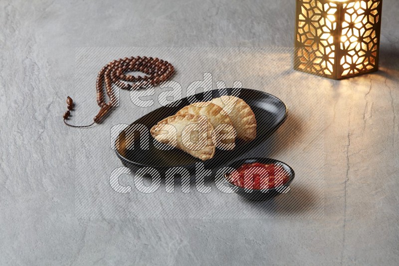 Three fried sambosas in an oval shaped black plate and a red sauce in a black round ramekin with a brown misbaha and a golden lantern on the side on a gray background