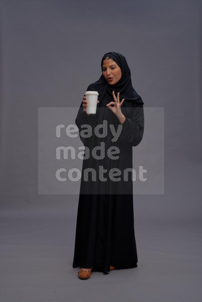 Saudi woman wearing Abaya standing holding paper cup on gray background