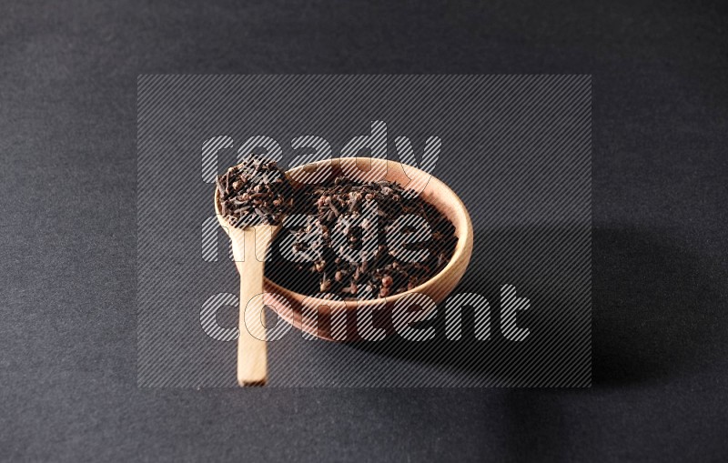 A wooden bowl and a wooden spoon full of cloves on a black flooring