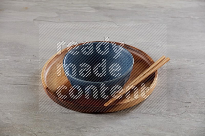 Blue pottery round bowl on top of brown wood round plate and wood chopsticks, on grey textured countertop