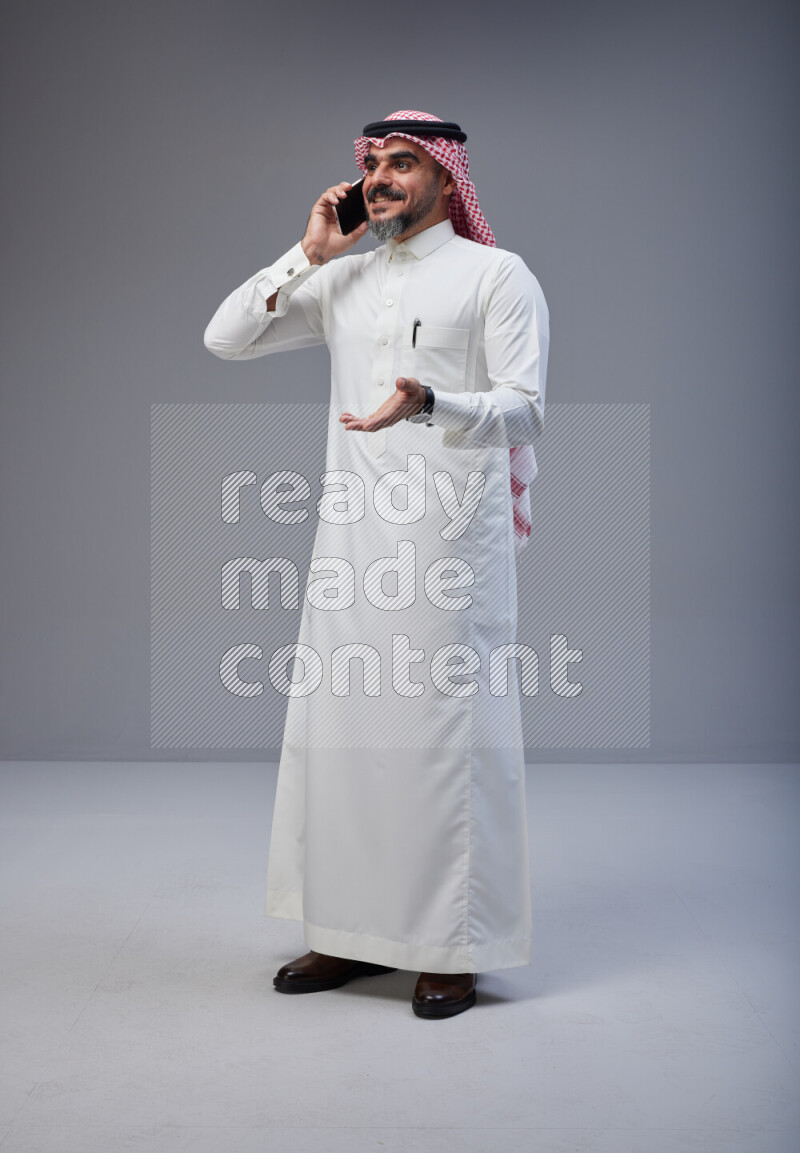 Saudi man Wearing Thob and red Shomag standing talking on phone on Gray background