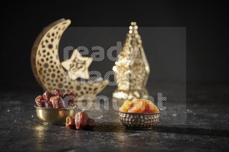 Dates in a metal bowl with dried apricot beside golden lanterns in a dark setup