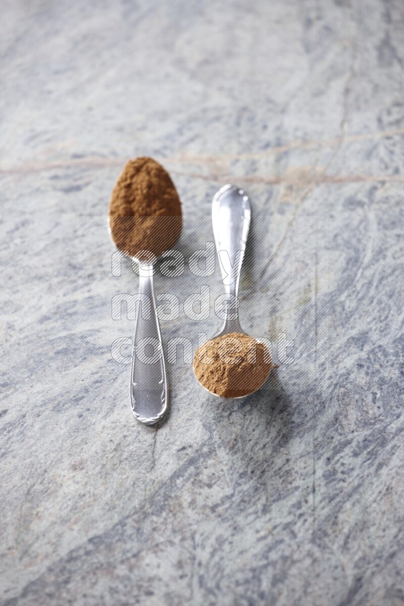 Two metal spoons full of cinnamon powder on grey marble background