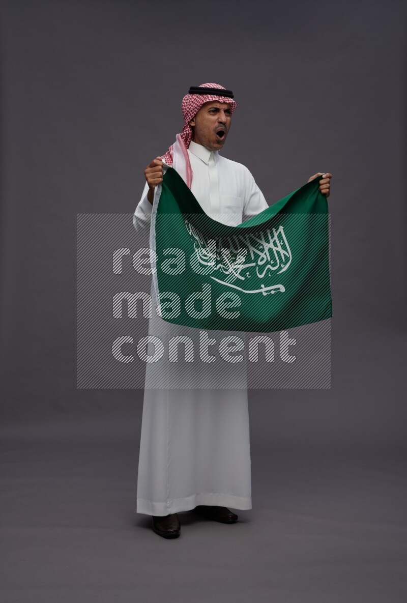Saudi man wearing thob and shomag standing holding Saudi flag on gray background