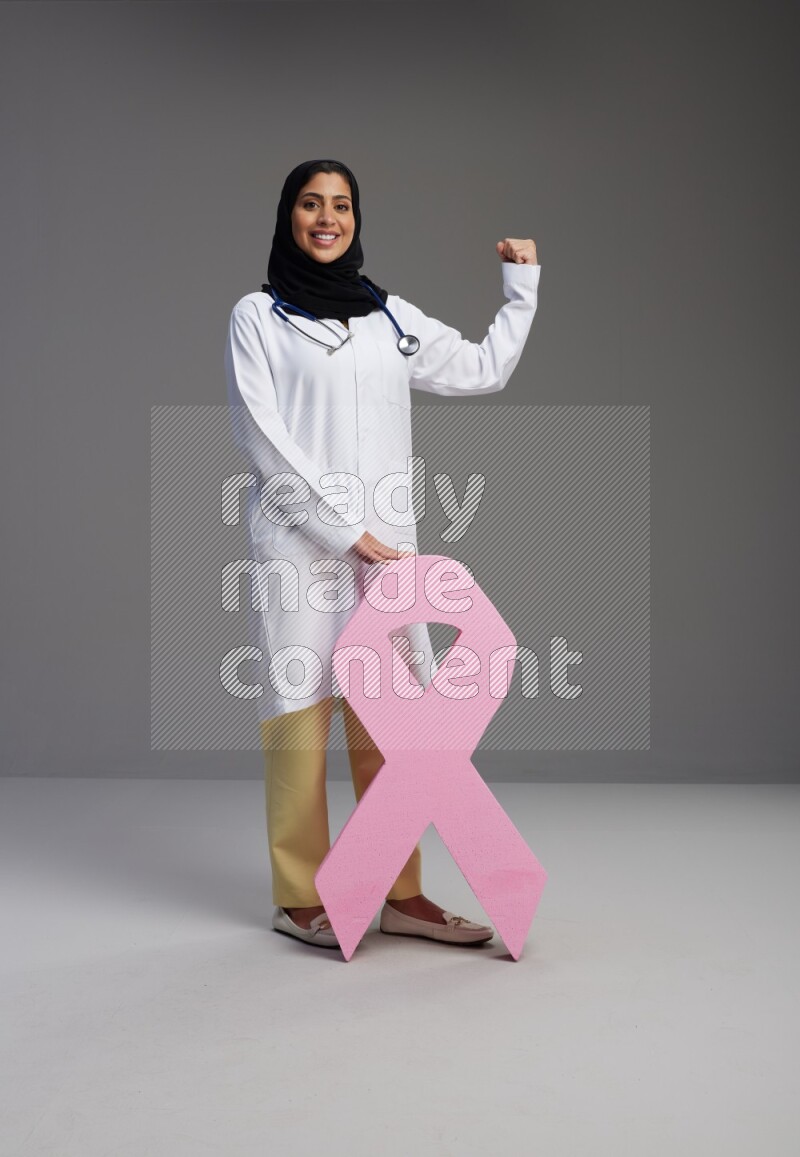 Saudi woman wearing lab coat with stethoscope standing holding awareness ribbon symbol standing on Gray background