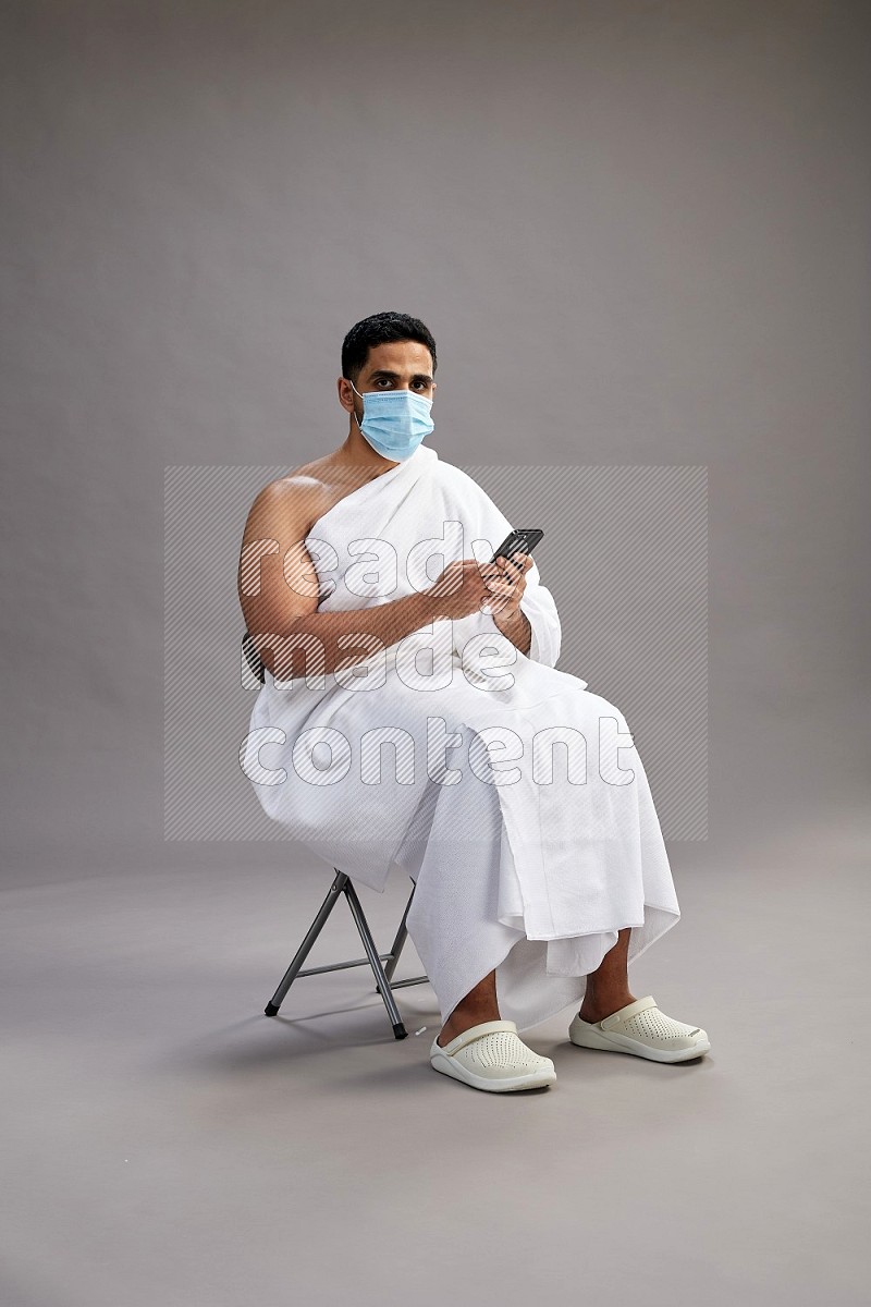 A man wearing Ehram with face mask sitting on chair texting on phone on gray background