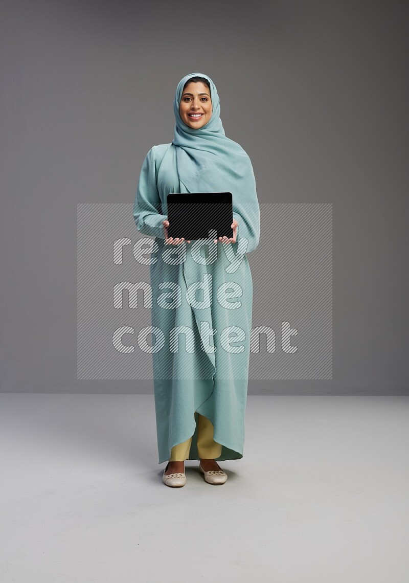 Saudi Woman wearing Abaya standing showing tablet to camera on Gray background