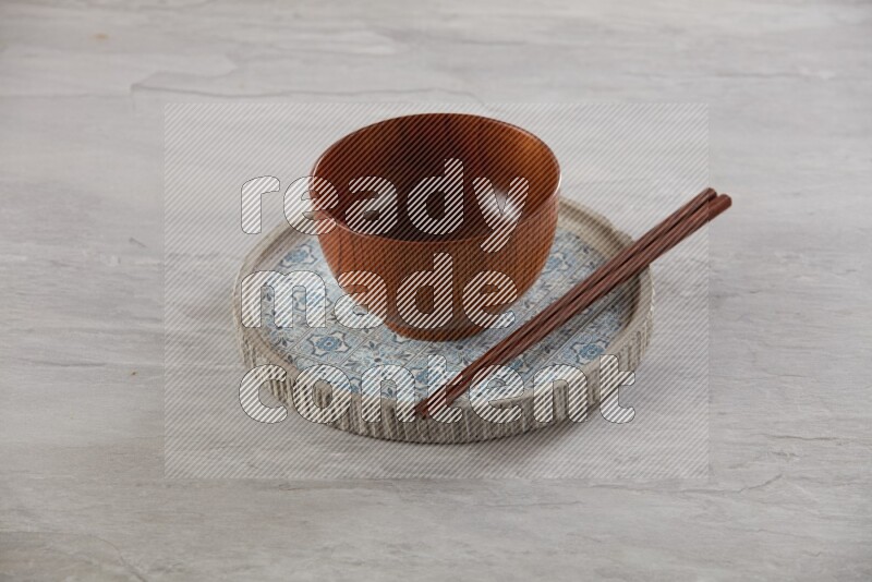 brown wood round bowl on top of multi color round ceramic plate and chopsticks, on grey textured countertop