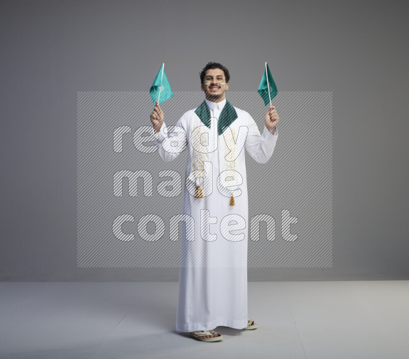 A Saudi man standing wearing thob and saudi flag scarf with face painting holding small Saudi flag on gray background