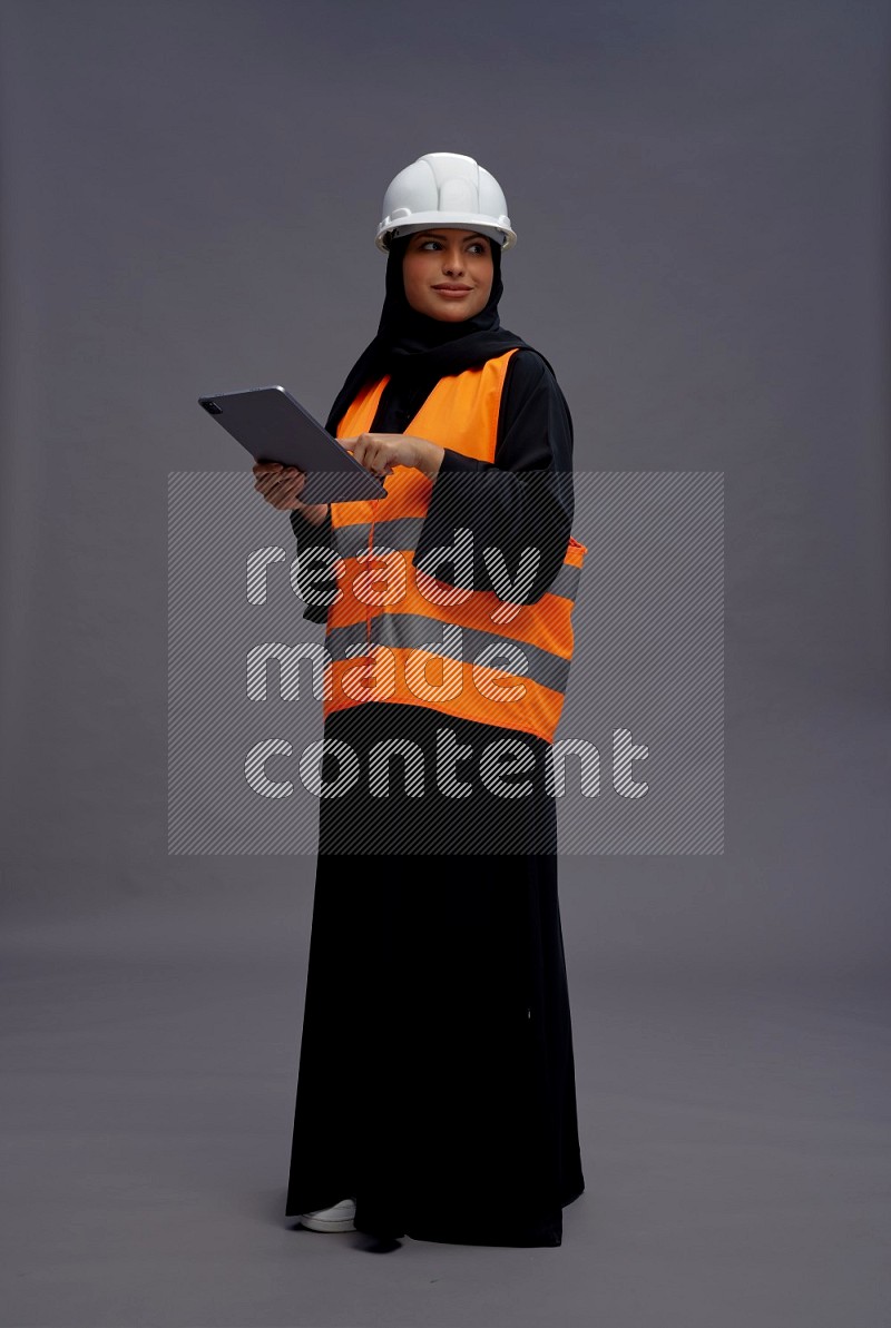 Saudi woman wearing Abaya with engineer vest standing working on tablet on gray background