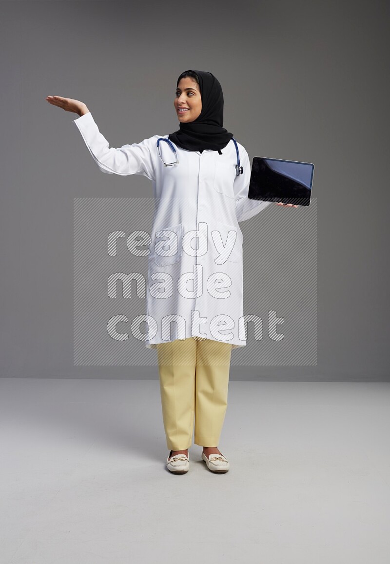 Saudi woman wearing lab coat with stethoscope standing showing tablet to camera with sign in the back on Gray background