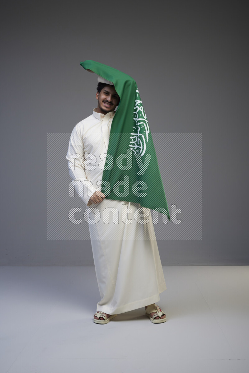 A saudi man standing wearing thob holding big saudi flag on gray background