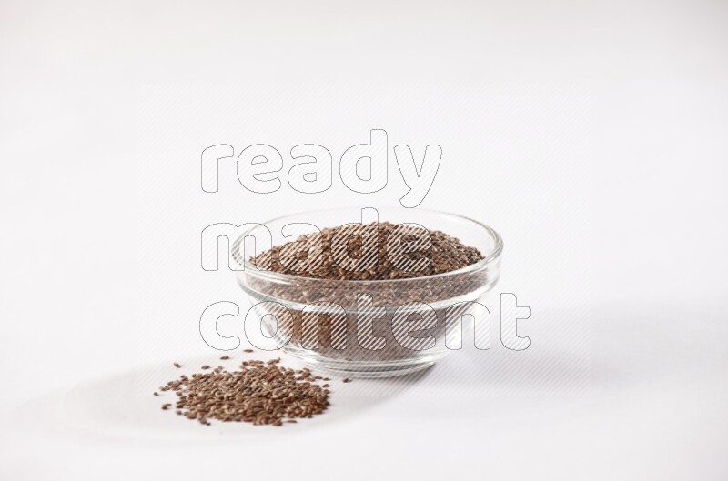 A glass bowl full of flax seeds surrounded by flax seeds on a white flooring