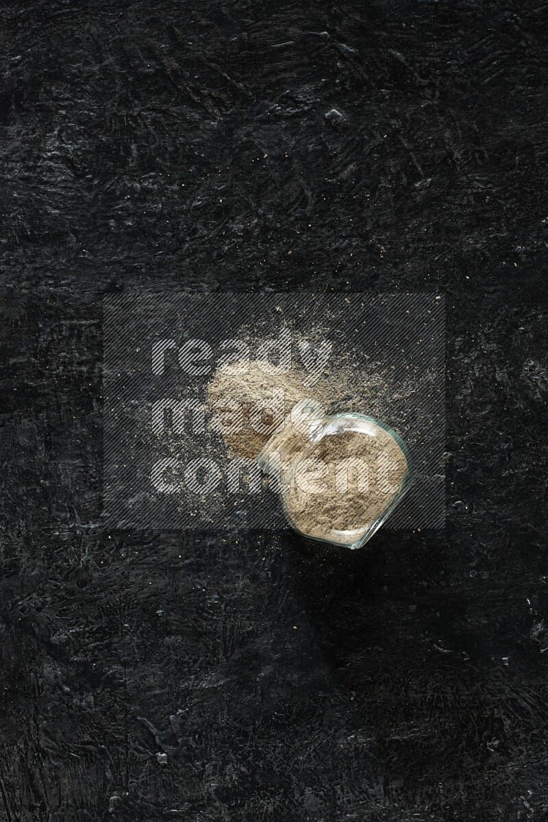 A flipped glass spice jar full of cardamom powder on textured black flooring