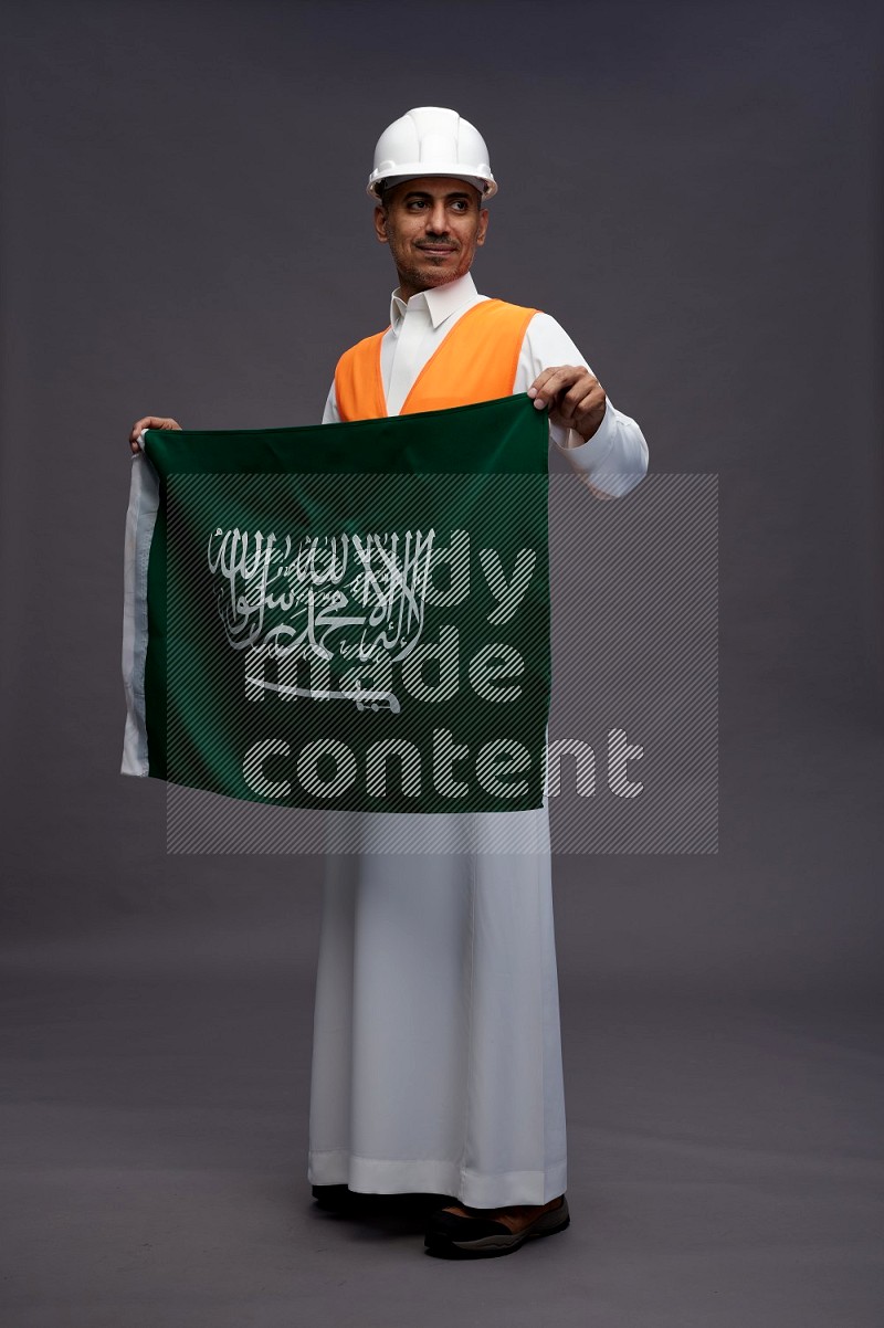 Saudi man wearing thob with engineer vest standing holding Saudi flag on gray background