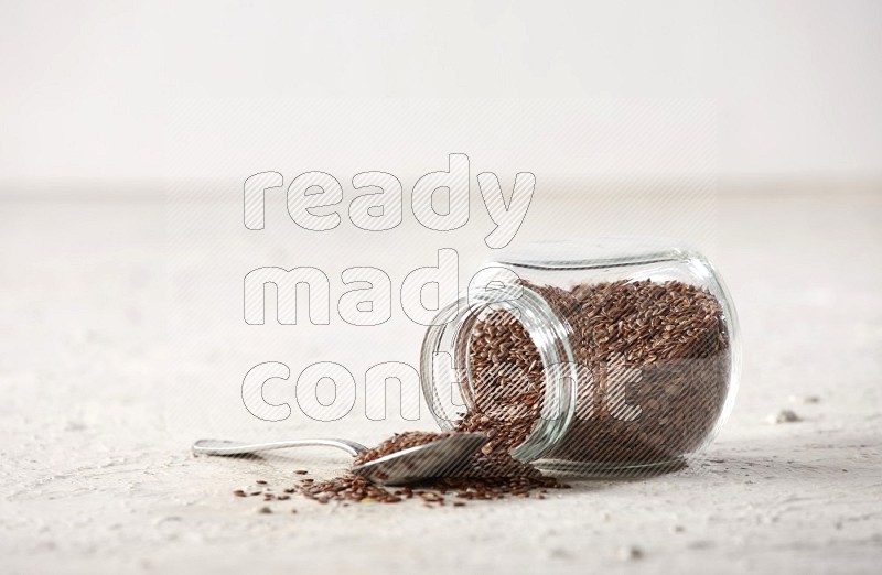 A glass spice jar full of flax flipped and a metal spoon full of the seeds on a textured white flooring in different angles