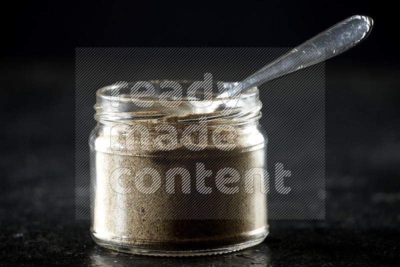 A glass jar and metal spoon full of cardamom powder on textured black flooring