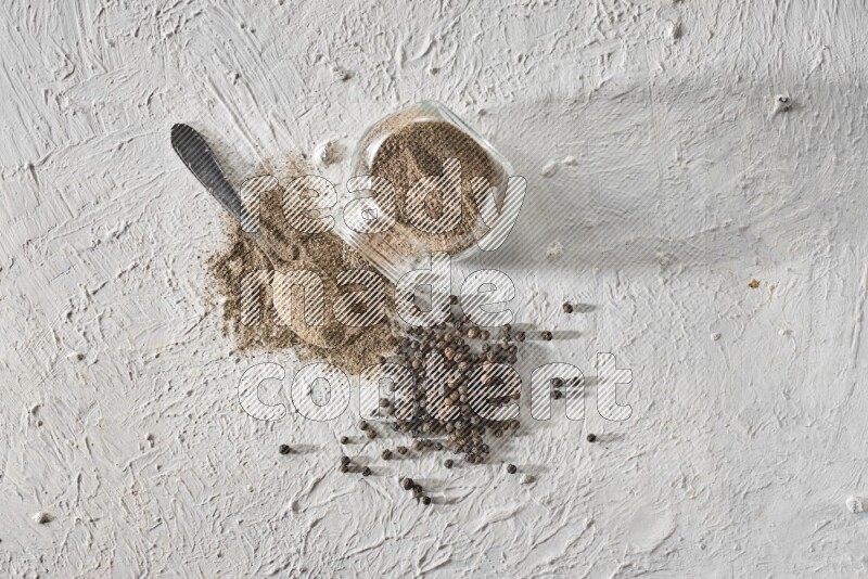 Flipped glass spice jar full of black pepper powder with a metal spoon and black pepper beads spread on a textured white flooring