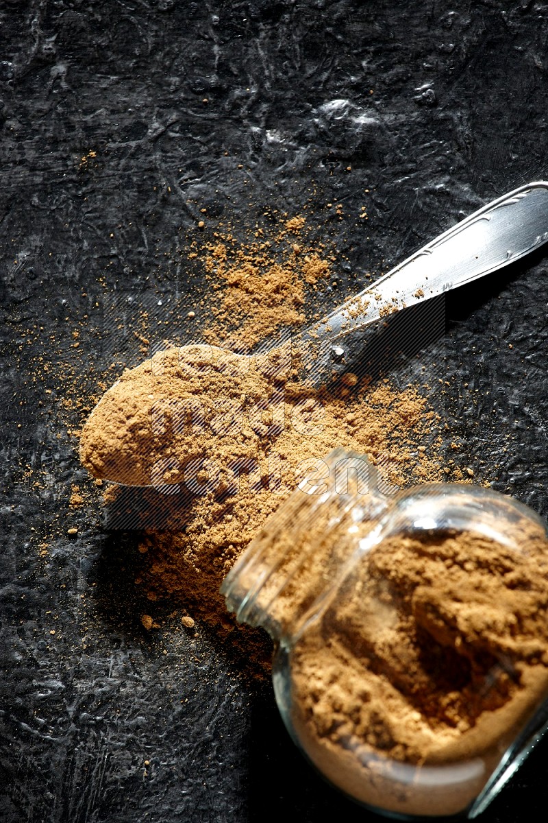 A flipped glass spice jar and a metal spoon full of allspice powder and powder spilled out of it on a textured black flooring