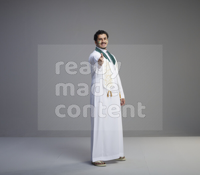 A Saudi man standing wearing thob and Saudi flag scarf with face painting on gray background