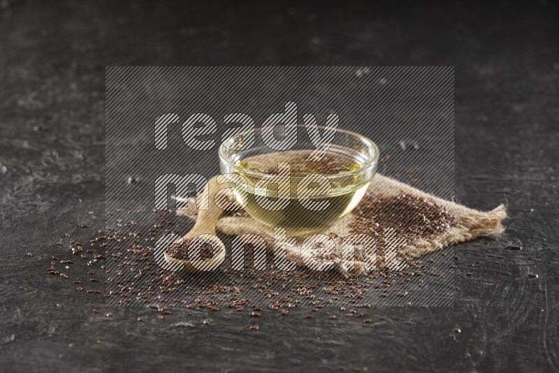 A glass bowl full of flaxseeds oil and wooden spoon full of flaxseeds with seeds spread on burlap fabric on a textured black flooring