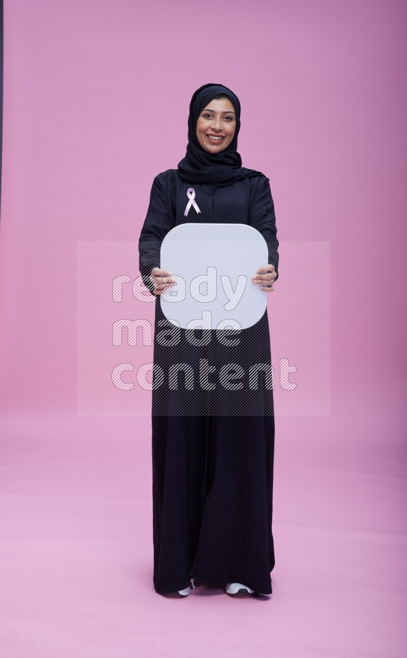 Saudi woman wearing pink ribbon on Abaya standing holding social media signs on pink background