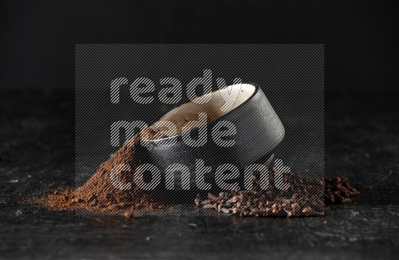 A black pottery bowl full of cloves powder spilled out of it with whole grains on a textured black flooring