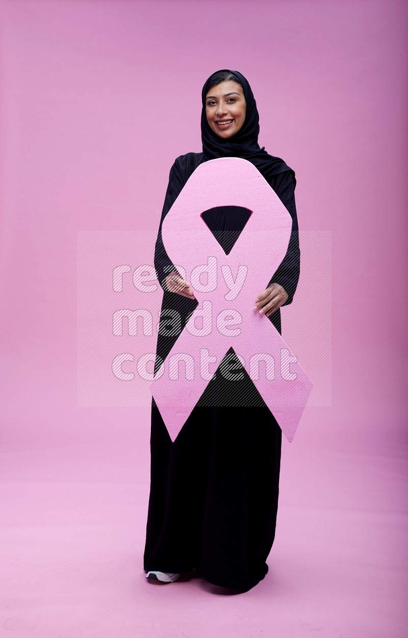 Saudi woman wearing pink ribbon on abaya standing holding awareness ribbon on plain pink background