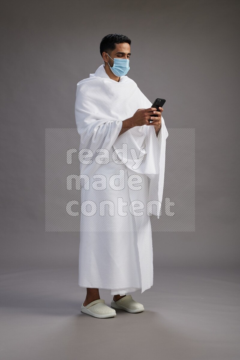 A man wearing Ehram with face mask Standing texting on phone on gray background