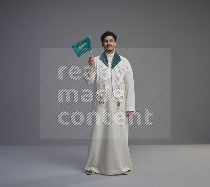 A Saudi man standing wearing thob and saudi flag scarf and holding small saudi flag on gray background
