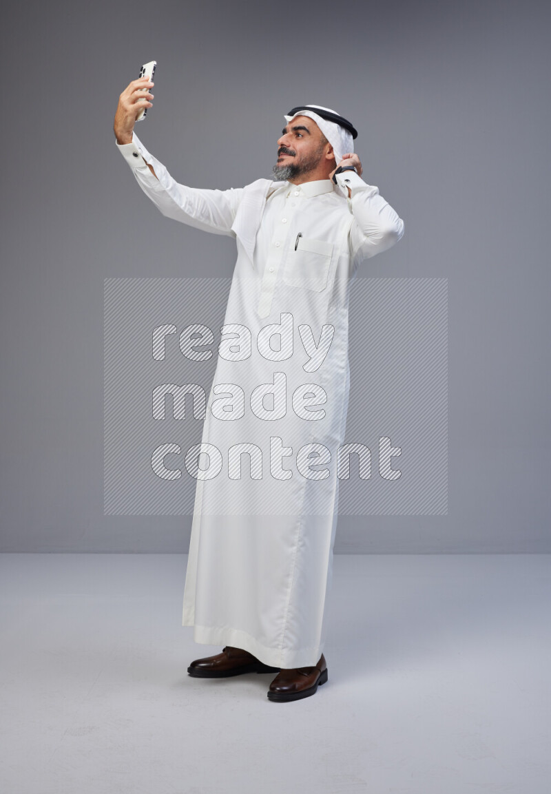 Saudi man Wearing Thob and white Shomag standing taking selfie on Gray background