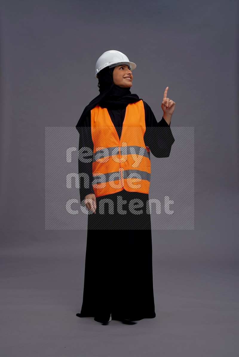 Saudi woman wearing Abaya with engineer vest standing interacting with the camera on gray background