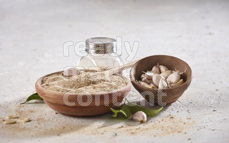 A wooden bowl with a spoon in it and glass spice jar, all full of garlic powder and a wooden bowl full of garlic cloves on a white flooring