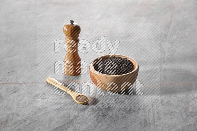 A wooden bowl full of black pepper and a wooden spoon full of black pepper powder and a wooden grinder on a marble flooring