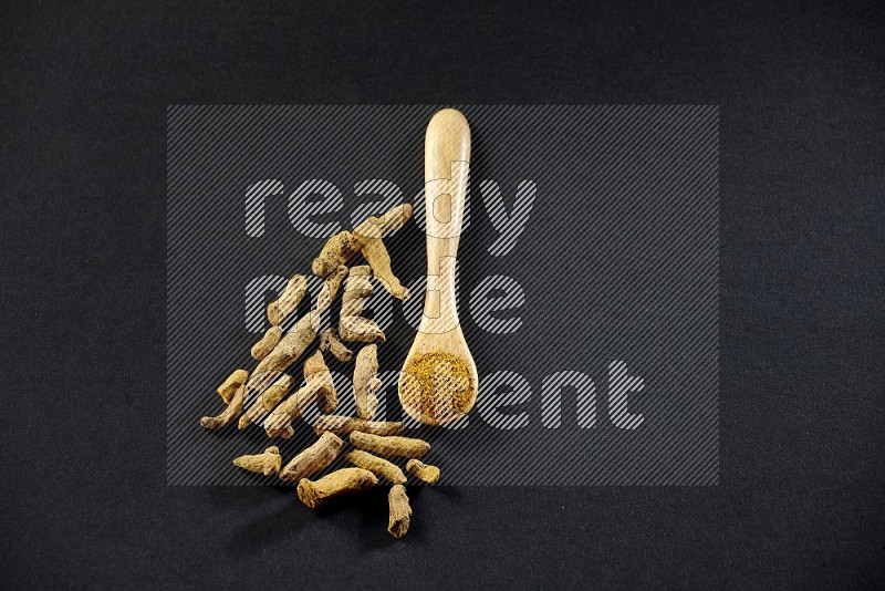 A wooden spoon full of turmeric powder with dried turmeric fingers on black flooring