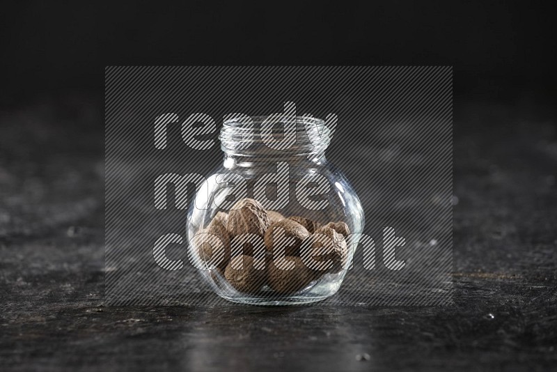 A glass spice jar full of nutmeg on a textured black flooring in different angles