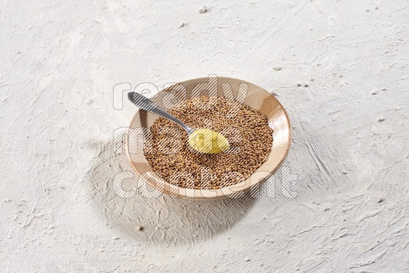 A beige pottery plate filled with mustard seeds with a metal spoon full of mustard paste in it on a textured white flooring