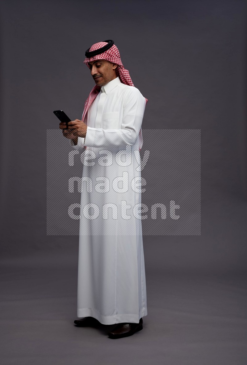 Saudi man wearing thob and shomag standing texting on phone on gray background