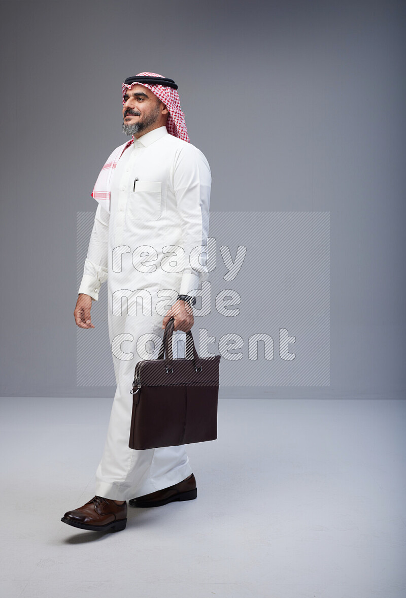 Saudi man Wearing Thob and red Shomag standing holding bag on Gray background