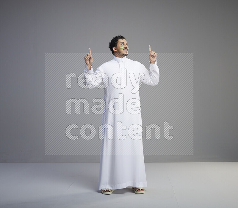 A Saudi man standing wearing thob with face painting interacting with the camera on gray background